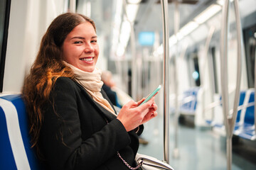 A cheerful young woman engaged in using her smartphone while seated on a subway train, fully...