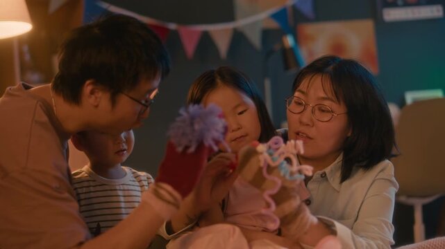 Happy modern Asian family with two little children spending evening time together in cozy warm lit bedroom, parents wearing sock puppets while entertaining kids before going to bed