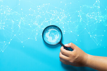 Global network connection and digital search concept. Hand holding a magnifying glass focusing on a world globe icon with abstract data lines, symbolizing technology, internet, and future analysis.