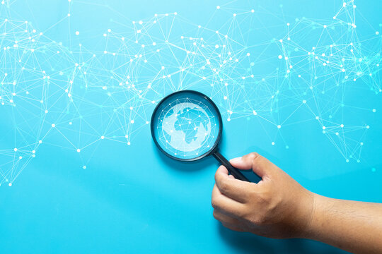 Global network connection and digital search concept. Hand holding a magnifying glass focusing on a world globe icon with abstract data lines, symbolizing technology, internet, and future analysis.