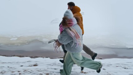 Happy Caucasian family of three in warm winter clothes holding hands as running along snowy path near frozen lake surrounded by forest on cold day