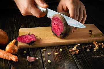 Hands slice a deep purple beet on a wooden board. Surrounding vegetables include garlic, carrots, and dried herbs, creating a cozy kitchen atmosphere