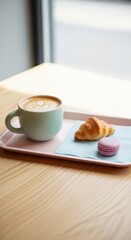 Lightly colored ceramic mug filled with coffee showing latte art rests on a tray with baked goods beside a bright window