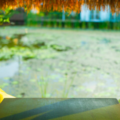 Wooden table with blurred natural lotus pond background under thatched roof, perfect for tropical product display, eco-friendly branding, or relaxing nature-themed designs.
