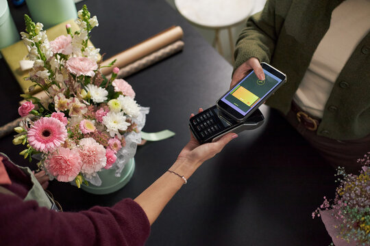 Caucasian young adult woman holding bouquet of assorted flowers while making contactless payment with smartphone at counter, florist processing transaction using card reader