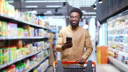 African american male buyer customer checking shopping list using smartphone, purchasing in grocery store in supermarket. Black man shopper pick goods use mobile phone in hypermarket or food market - Powered by Adobe