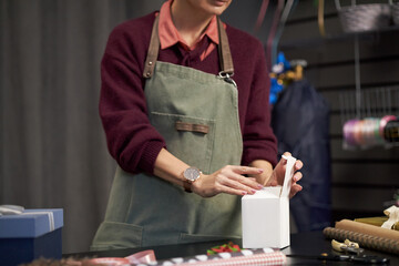 Caucasian young adult woman arranging ribbon on gift box at flower shop counter, hands visible tying bow, various wrapping supplies and floral accessories in background