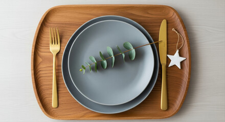 Wooden charger with gray plate and brass cutlery. Eucalyptus sprig and small white ornament completing placement.  Nordic style Christmas table setting, wood, gray, gold accents
