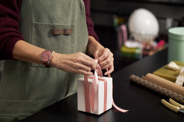 Caucasian young adult woman tying pink ribbon on gift box at table, hands visible arranging bow, preparing present in workspace with wrapping supplies and decorative items nearby