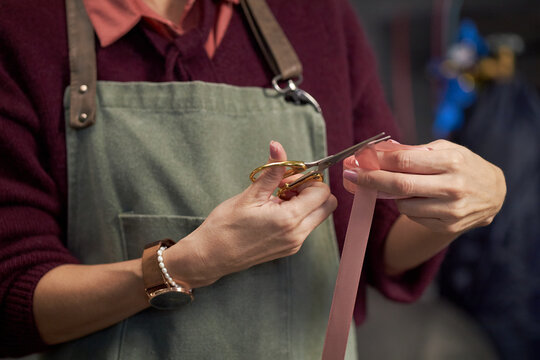 Caucasian middle aged woman wearing apron holding scissors and cutting pink ribbon with both hands, focusing on precise work, hands and torso visible, no face shown - Powered by Adobe