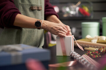 Caucasian young adult woman wrapping gift box with pink ribbon on table, surrounded by rolls of...