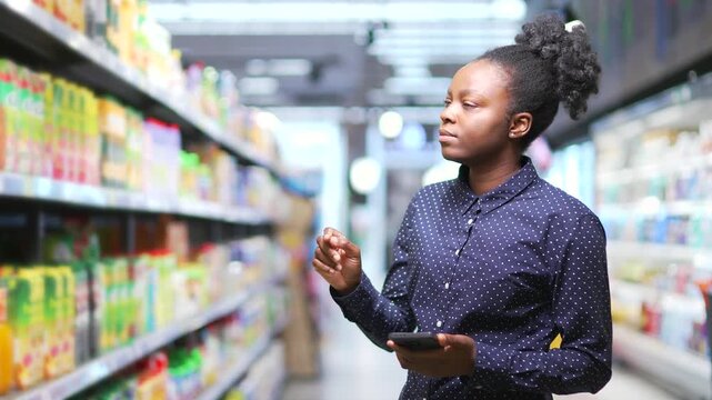 Young african american woman choosing products stands in a grocery store in a supermarket. Thoughtful woman shopper checking shopping list using smartphone looking at shelves. Browses through products