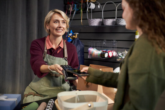 Caucasian middle aged woman wearing apron smiling, while handing payment terminal to young adult Caucasian woman making contactless payment at flower shop counter with gift bags visible