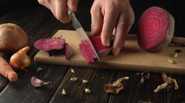 A person uses a sharp knife to cut a fresh purple beet into neat slices on a wooden cutting board. Surrounding ingredients include carrots, garlic, and dried spices.