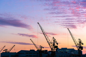 Fototapeta premium Silhouettes of construction cranes and industrial machinery at a port or shipyard against a beautiful sunset sky with purple and pink clouds.