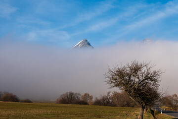 High Tatras peaks emerging from a thick inversion fog above fields and trees under a clear blue sky.