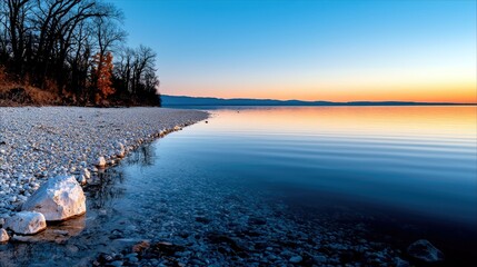 A serene sunrise over a calm lake, with a rocky shore and bare trees reflecting in the water.