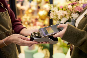 Caucasian middle aged woman holding payment terminal while Caucasian young adult woman making contactless payment with smartphone in flower shop with colorful floral background