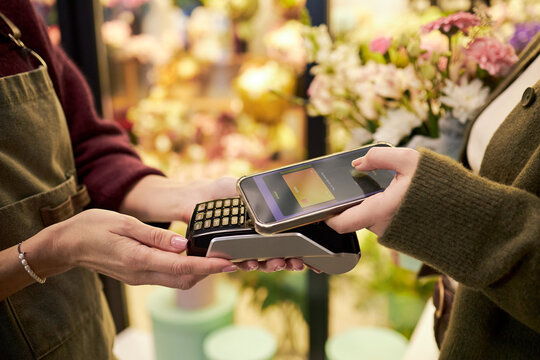 Caucasian middle aged woman holding payment terminal while Caucasian young adult woman making contactless payment with smartphone in flower shop with colorful floral background