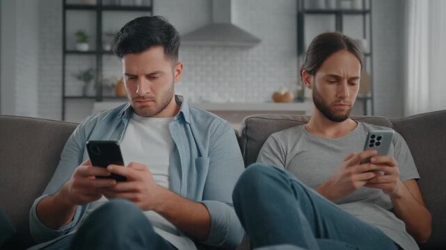 Two men sit apart on a sofa, deeply engrossed in their smartphones, illustrating a modern communication breakdown and digital distraction in a relationship