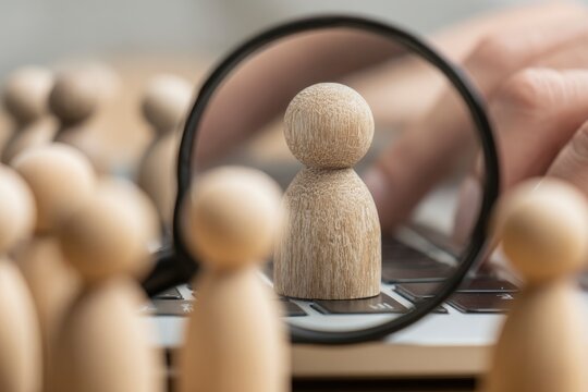 Wooden figures are placed on a laptop one is highlighted by a magnifying glass and a finger hovers over the keyboard