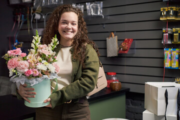 Young adult Caucasian woman smiling while holding large bouquet of assorted flowers in pastel bucket, standing in flower shop with gift boxes and floral supplies visible in background