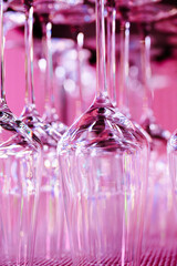 rows of clean upside-down wine glasses arranged on a shelf and illuminated with soft pink lighting. The reflective glass surface creates a modern, stylish atmosphere suitable for themes related to res