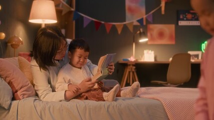 Cozy evening scene of Asian family at home with mother reading to her son in background while daughter playing with basketball in warmly lit, comfortable room