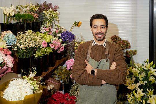 Young adult Black man standing with arms crossed in front of colorful flower arrangements in florist shop, smiling confidently, surrounded by various fresh blooming flowers