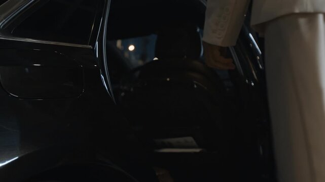 Cropped shot of successful woman getting into back seat of business class car while personal driver in white gloves opening door for client in night city street