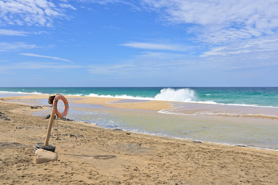 Lifebuoy standing on a sandy coast at El Cotillo beach, Playa del Castillo, Fuerteventura. Orange rescue ring on the sandy beach with breaking sea waves under blue sky.