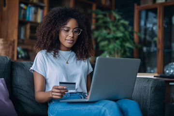 Online Shopping Experience: A woman, wearing glasses, sits on a comfortable couch, using a laptop and a credit card, symbolizing the convenience of online shopping.