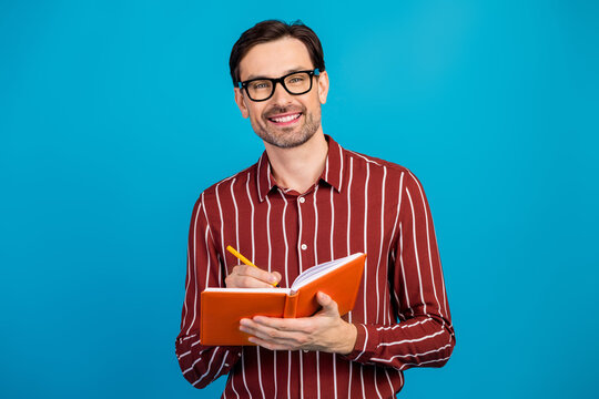 Young handsome man with striped shirt writing in orange notebook against blue background