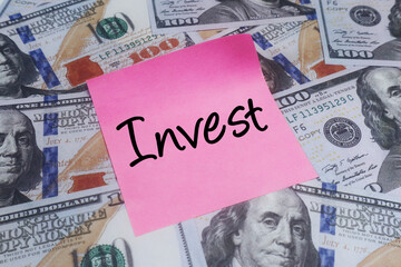 Pink sticky note with Invest handwritten, placed on a backdrop of hundred dollar bills, symbolizing financial planning, wealth building, and future economic growth strategies