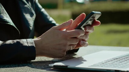 Woman scrolling phone next to laptop outdoors. Female hands navigating smartphone screen under sunlight. Unrecognizable user browsing social media beside portable computer. Remote worker using mobile - Powered by Adobe