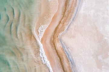 This scene reveals a beautiful coastal landscape seen from above, showcasing clear shallow waters meeting sandy shores. Subtle waves gently lap against the beach, revealing intricate patterns
