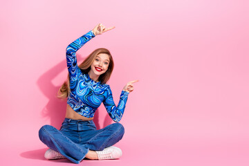 Glamorous young woman in blue printed top sits on pink background smiling and pointing
