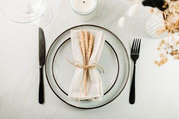 Elegant minimal table setting with ceramic plates, black cutlery and a linen napkin decorated with wheat stems, styled in a soft natural light.