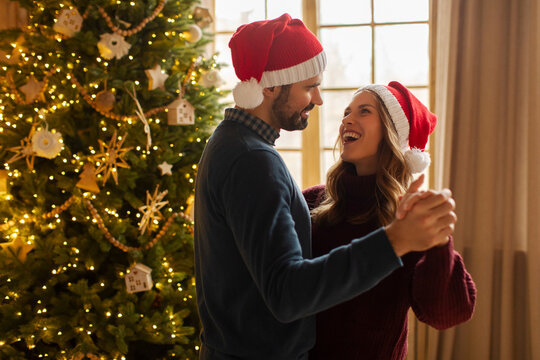 Happy couple dancing near decorated Christmas tree, wearing festive Santa hats, celebrating New Year at home, free space