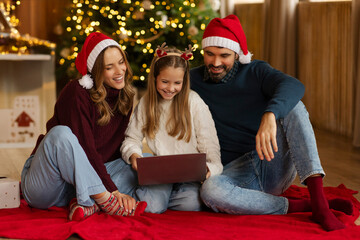Family sitting on blanket in front of decorated Christmas tree, sharing joyful moments while...