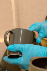 close-up of female hands wearing protective gloves while washing dishes under running water. A simple everyday household scene that conveys cleanliness, care, and routine home maintenance.