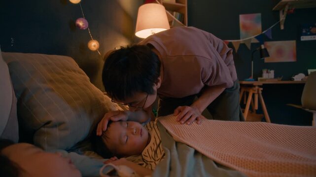 Caring Asian father adjusting his child blanket and gently kissing them goodnight, sharing tender bedtime moment in cozy, warm bedroom at night