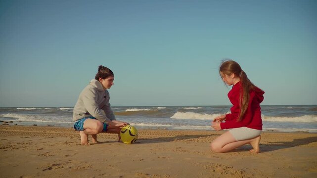 Teenagers sitting on sand playing ball together on sea shore. Children crouching on beach enjoying ball game. Boy and girl interacting with ball on seaside sand