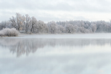 Frost-covered trees reflected in calm water with morning mist. Overcast sky and soft winter light.