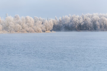 Winter forest covered in hoarfrost reflected in calm lake. Mist rises from water, soft morning light.