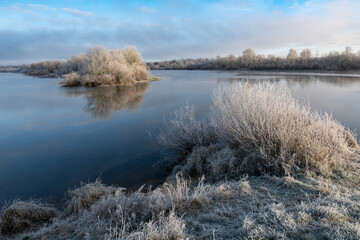 Island with frost-covered trees reflected in calm water. Foreground grasses and soft winter light