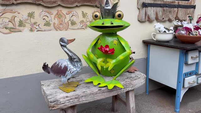 Close-up view of a charming souvenir shop entrance in Hallstatt, Austria. A funny metal frog king with a crown and a duck statue sit on a wooden bench. Next to them, an vintage blue stove is used as a