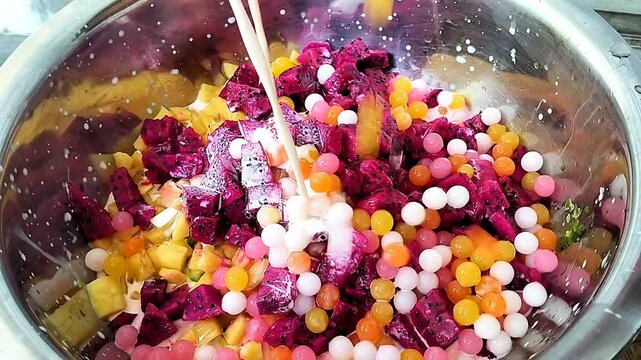 Preparation of a tropical fruit dessert: Coconut milk poured over mixed tropical fruit and colorful jelly pearls in a stainless steel container