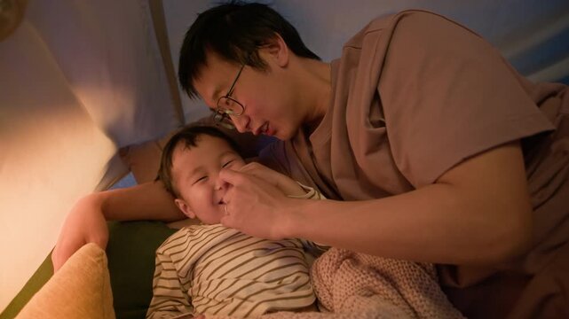 Asian father and little son lying together inside cozy teepee tent, sharing tender and loving moment in warm evening light, expressing affection and family bonding at home