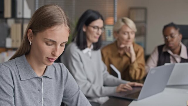 Tilt up shot of focused young Caucasian female employee drafting business project or graph on paper while her diverse female colleagues coworking in front of laptops in blurred background